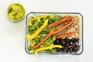 glass lunch container with quinoa, lettuce, black beans and red and yellow peppers. side of guacamole 