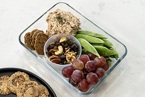 glass lunch container with grapes, pea pods, tuna salad, mixed nuts and crackers