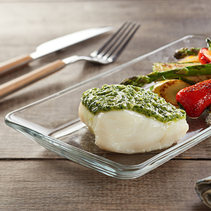 Fresh Pesto with Baked Wild Cod on a glass platter atop a wooden table with utensils nearby.