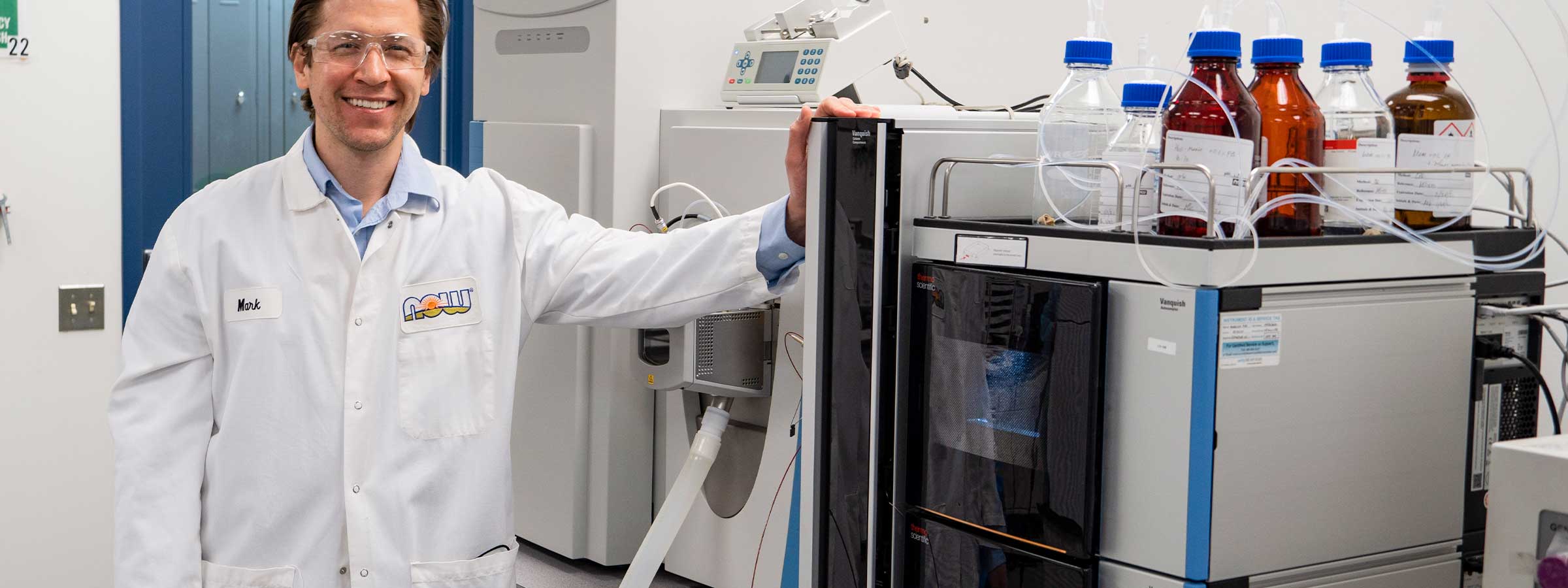 light skin male presenting person standing in a white lab coat next to a machine in a lab setting