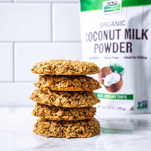 stack of five coconut oat cookies on a counter with bag of NOW Coconut Milk Powder behind it