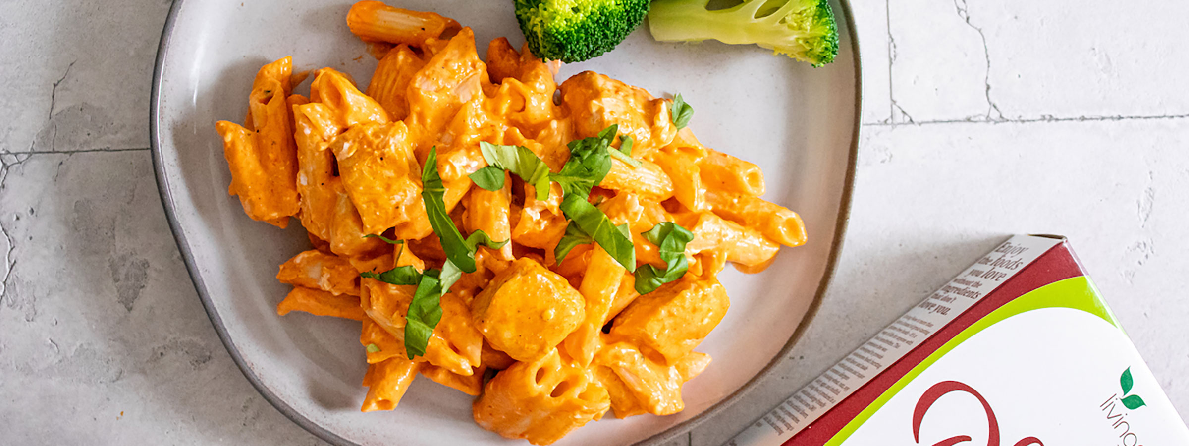 cooked NOW Gluten-free Penne Pasta on a plate covered with creamy yellow sauce and broccoli also on plate. Partial view of box of NOW Penne pasta.