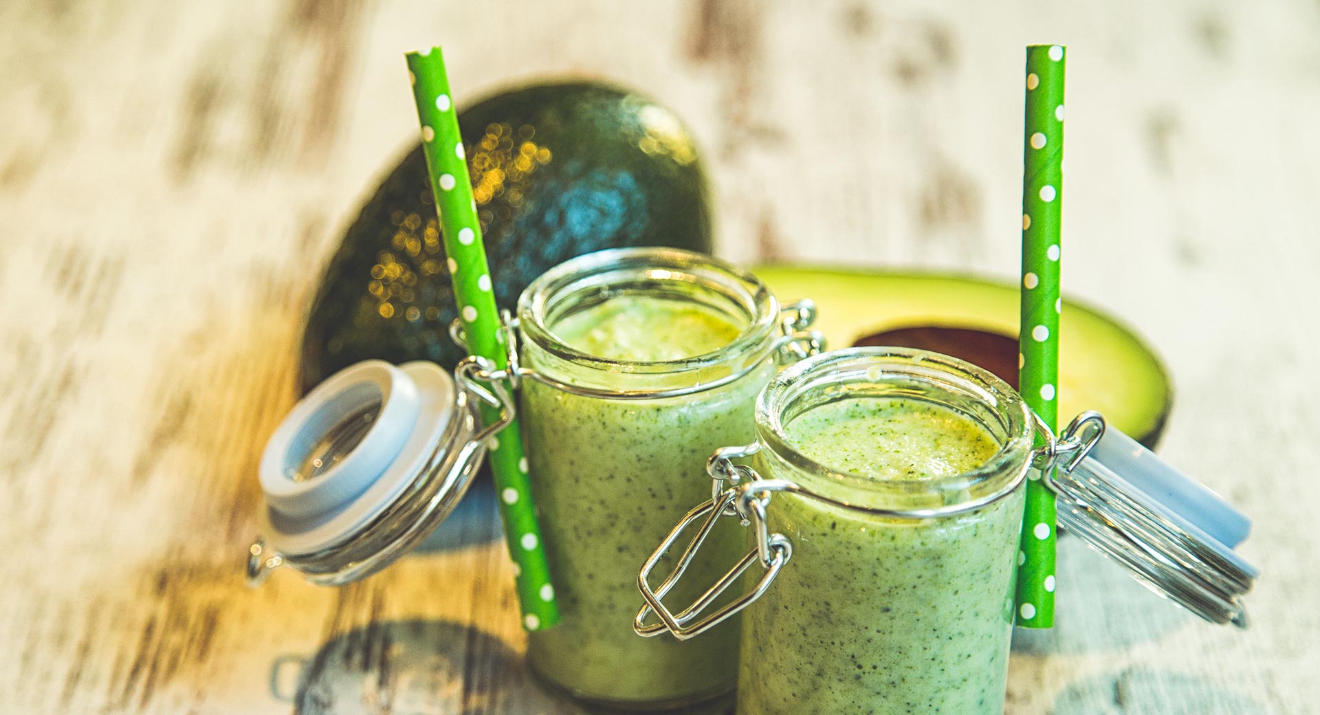 green smoothie in glass jars with green straws, avocado on counter behind glass