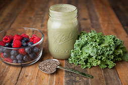 bowl of raspberries and blueberries beside a smoothie in a jar, and a bunch of kale