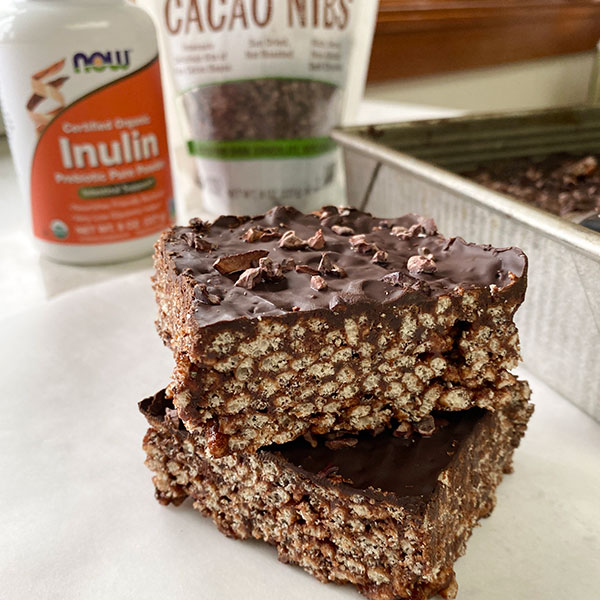 two dark chocolate brown rice crispy treat squares stacked on a white counter with a bottle of NOW Inulin and a package of NOW Cacao Nibs in the background