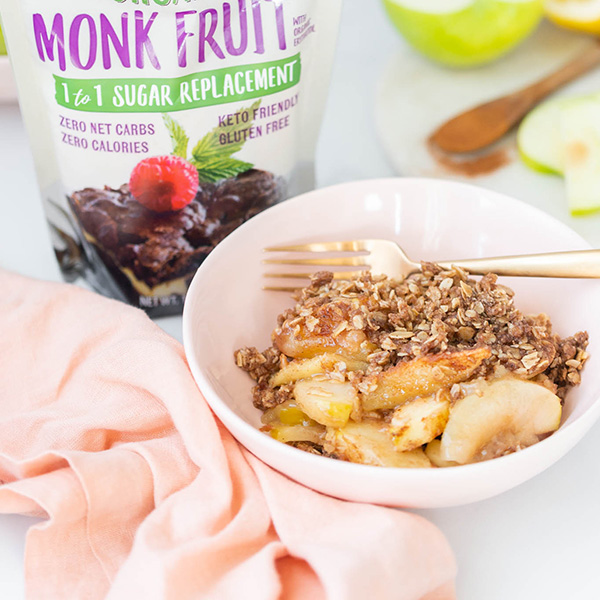 spiced apple crisp in a light pink bowl with a small scoop of ice cream on top and a bag of NOW Monk Fruit Sweetener to the left of the bowl