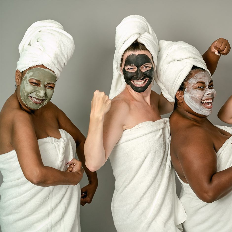 three female presenting persons with light and dark skin wearing white towels on their heads and body. All have face-masks in varying shads of light and dark green. 