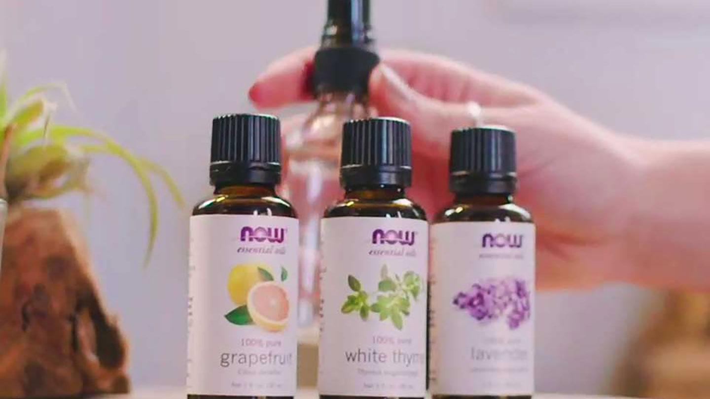 light skin hand grabbing an empty spray bottle with 3 bottles of NOW essential oils in front of it