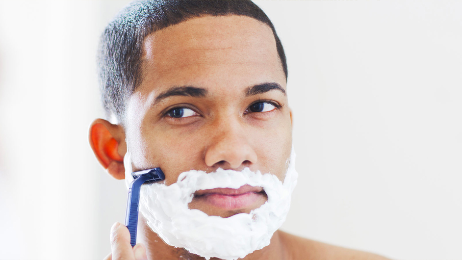 male presenting person with medium skin tone shaving with a blue razor and shaving cream on his face
