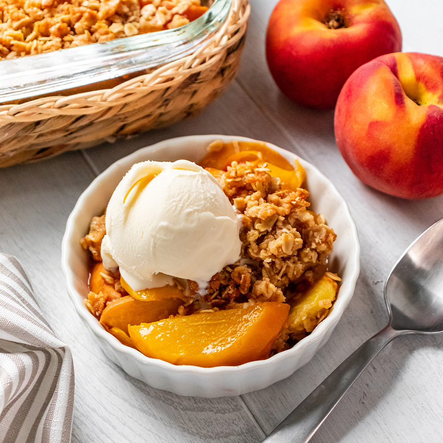 top view of bowl with peach crumble with an oat topping and a dollop of ice cream next to two peaches