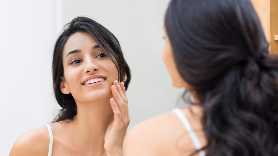 female presenting person with dark hair looking in a mirror touching her face
