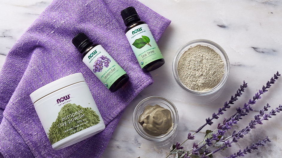 top view of NOW European Clay Powder, NOW Lavender Essential Oil, and NOW Tea Tree Essential Oil with a purple towel all on top of a marble table
