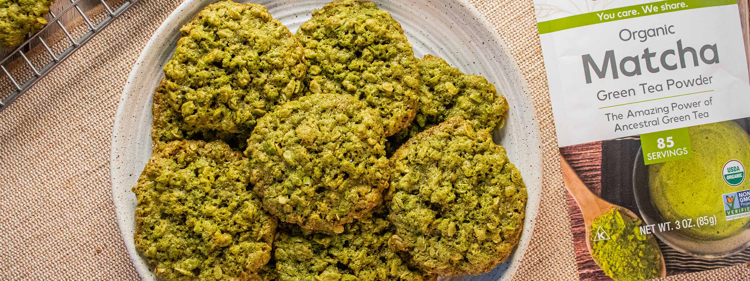 top view of a light brown surface containing a white plate with Chewy Matcha Oatmeal Cookies next to a bag of NOW Matcha tea