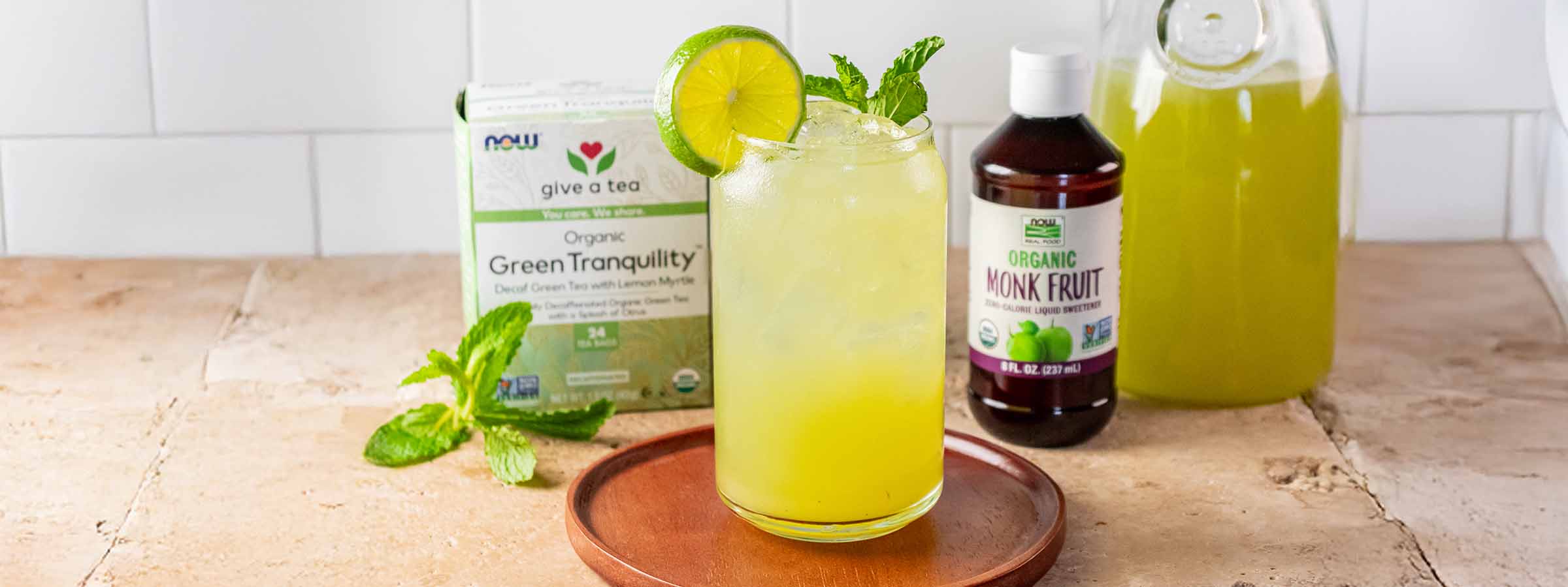 stone countertop with a glass of Citrus Cucumber and Mint Refresher and a box of NOW Green tea and NOW Monk Fruit Liquid
