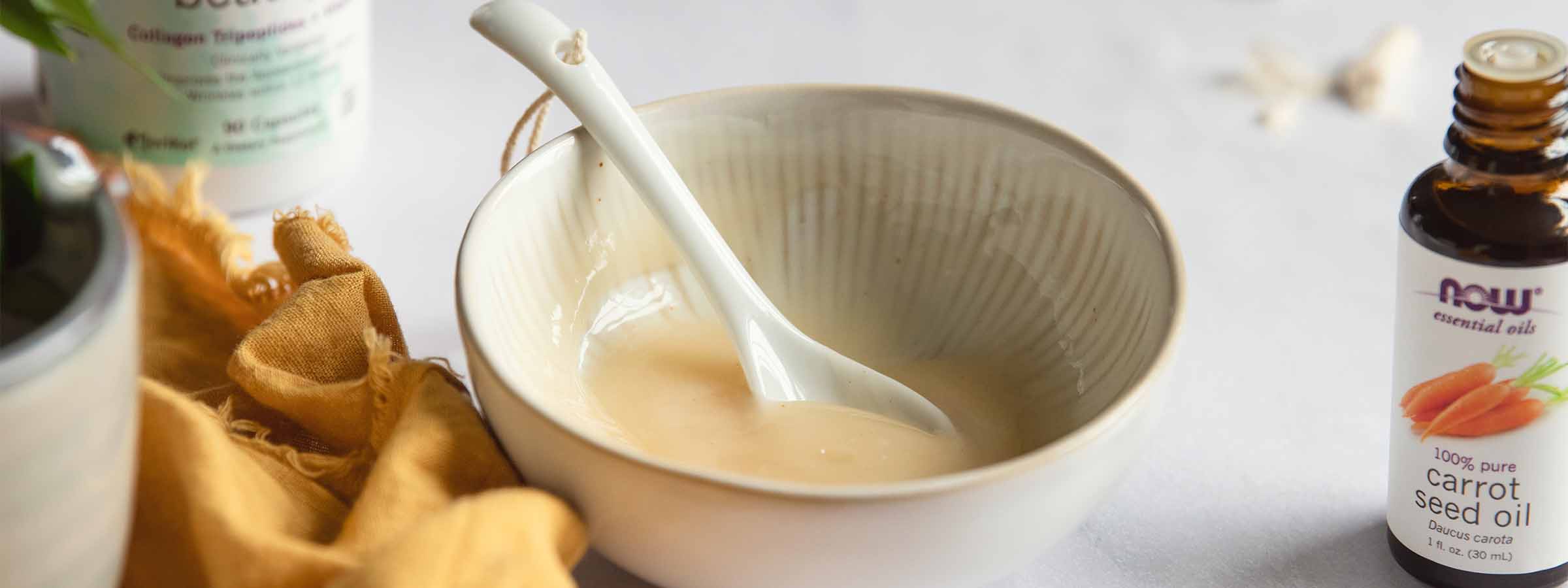 bowl of Cooling Collagen Face Mask next to NOW® Carrot Seed Essential Oil and a bottle of NOW® Solutions Aquatic Beauty+™ in the background