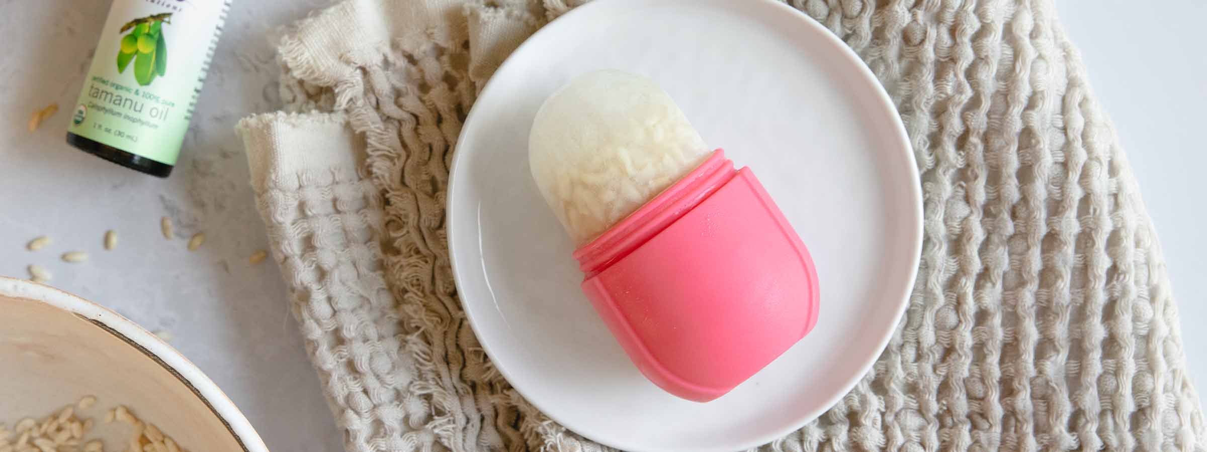 top view of Rice Water Recovery Ice Roller next to a bottle of NOW® Solutions Organic Tamanu Oil, all on a brown terry cloth