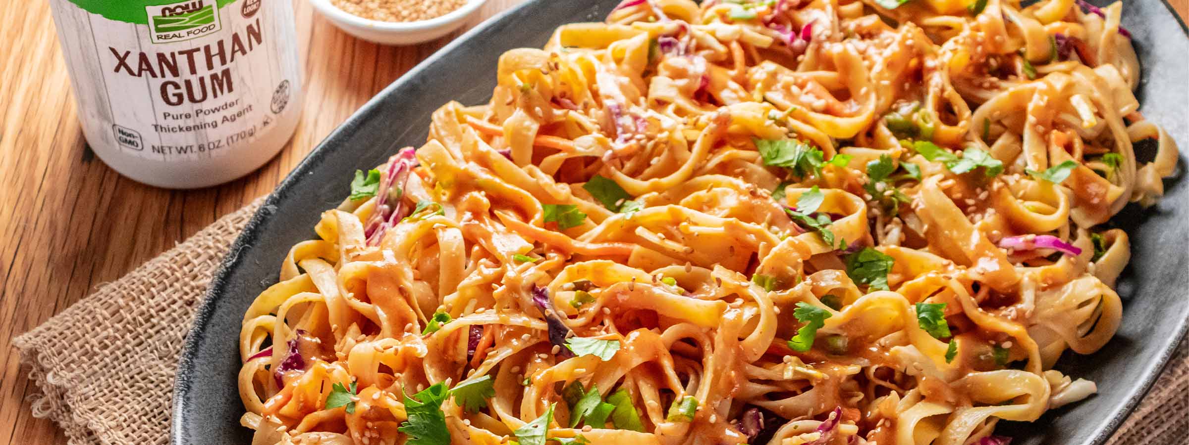 top view of oval black dish with Sesame Noodle Salad with Nutty Dressing inside it, next to NOW Xantham gum