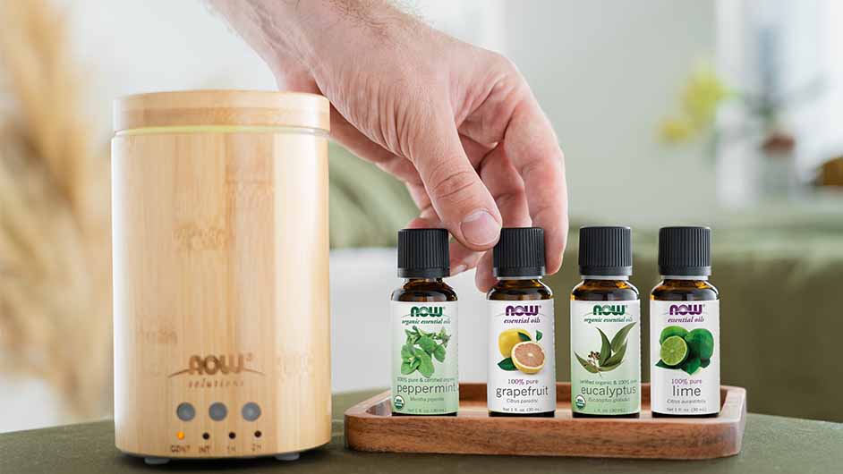 NOW Bamboo diffuser next to peppermint, grapefruit, eucalyptus, and lime essenital oils with light skin hand grabbing the grapefruit one.