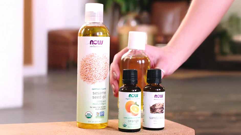 ingredients for detox massage oil including Sandalwood Oil blend, Orange Oil and an unamed carrier oil with a hand reaching in for a bottle of the mixed recipe