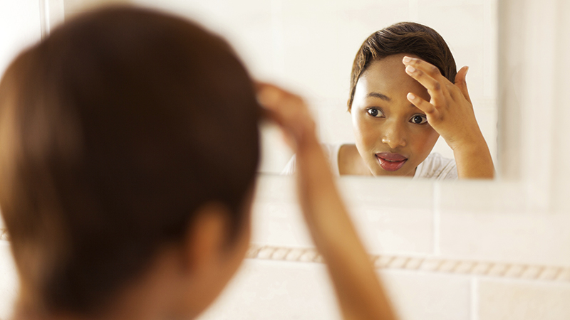 A female-presenting person with dark skin and short brown hair looking at themselves in the mirror