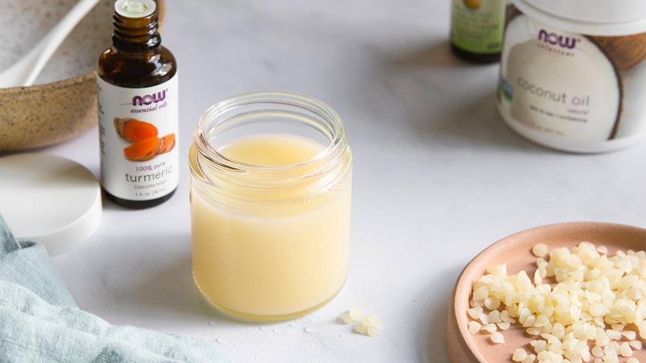 glass jar filled with Turmeric Oil Cleanser, Bottle of NOW Turmeric Essential Oil, NOW Solutions Coconut Oil, and beeswax pellets