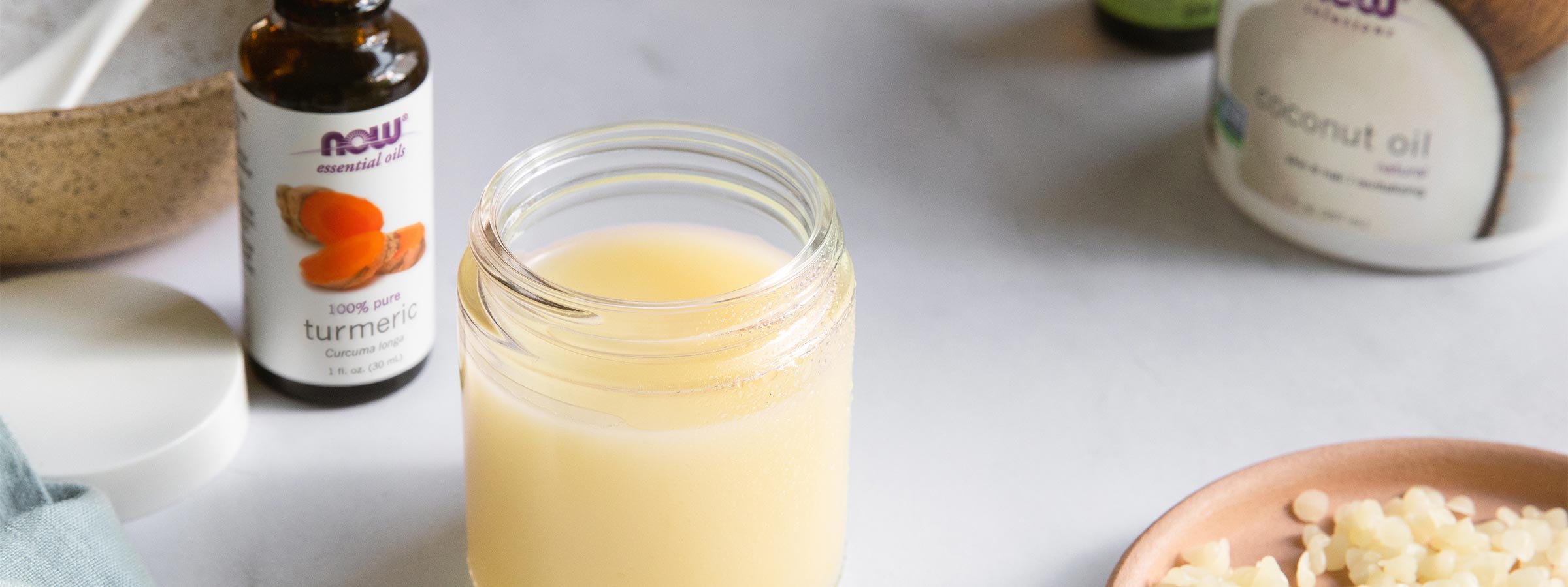 glass jar filled with Turmeric Oil Cleanser, Bottle of NOW Turmeric Essential Oil, NOW Solutions Coconut Oil, and beeswax pellets