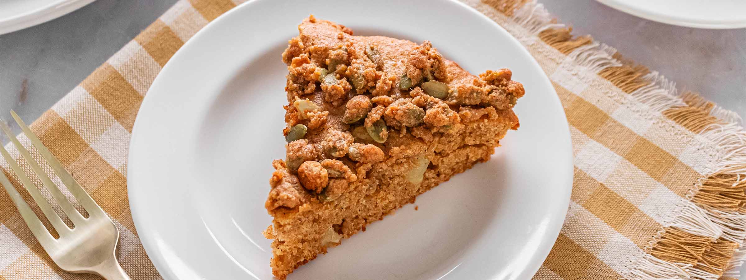 close up image of  Apple Spice Coffee Cake with Pumpkin Seed Crumble slice on white plate and checkered cloth