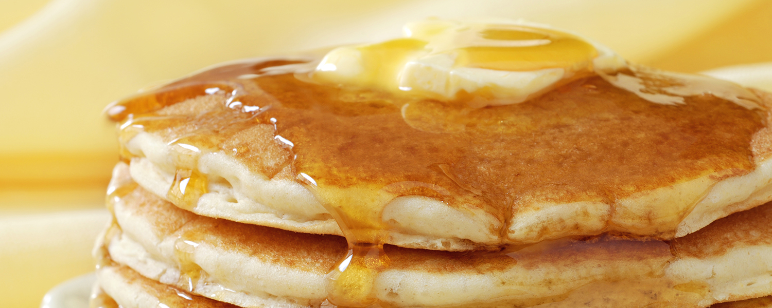 A stack of fluffy Buttermilk Pancakes doused in syrup with a dollop of butter melting on top.