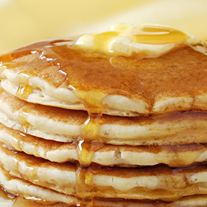 A stack of fluffy Buttermilk Pancakes doused in syrup with a dollop of butter melting on top.