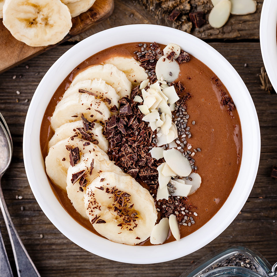white bowl of Chocolate smoothy with slices of bananas, chia seeds, coconut and chocolate shavings