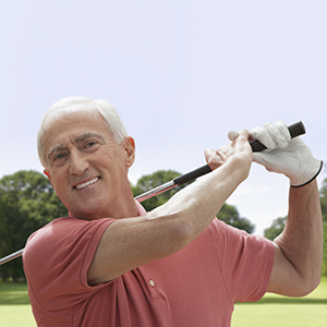 A male-presenting, light-skin person with grey hair smiles during a swing at an outdoor golf course.