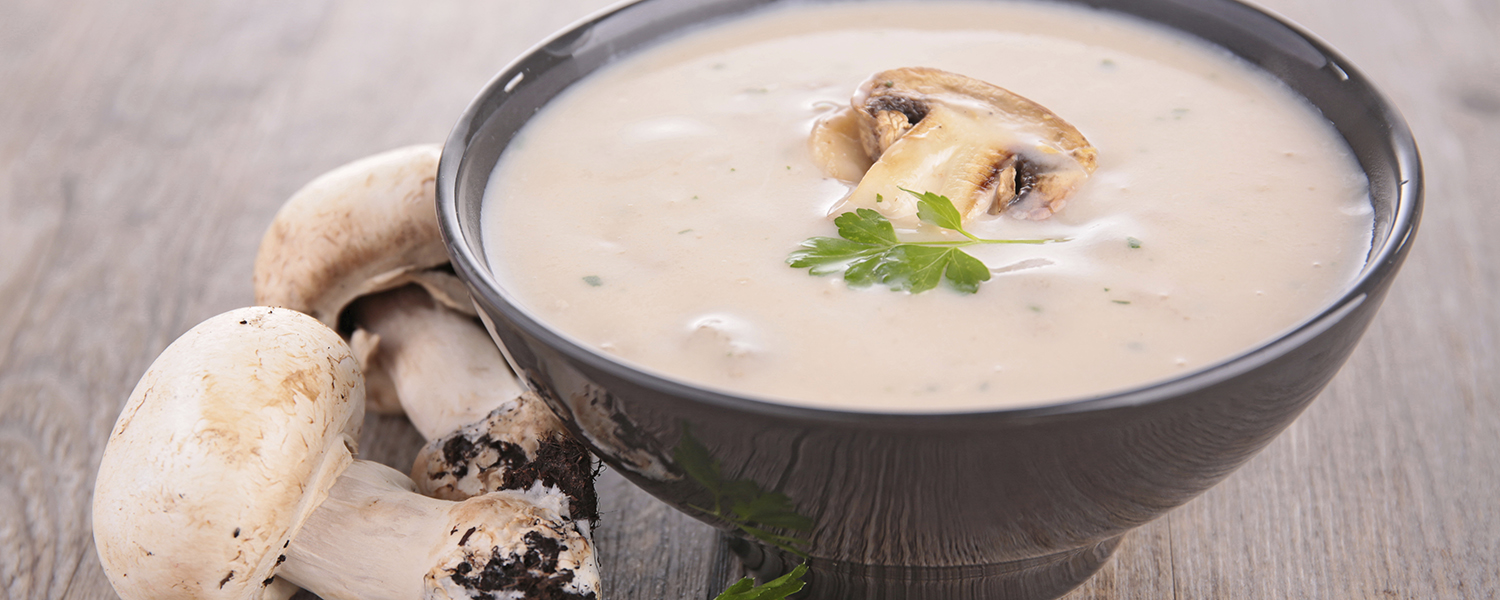 A grey soup bowl on a wooden table surrounded by whole mushrooms is full of Creamy Mushroom Soup