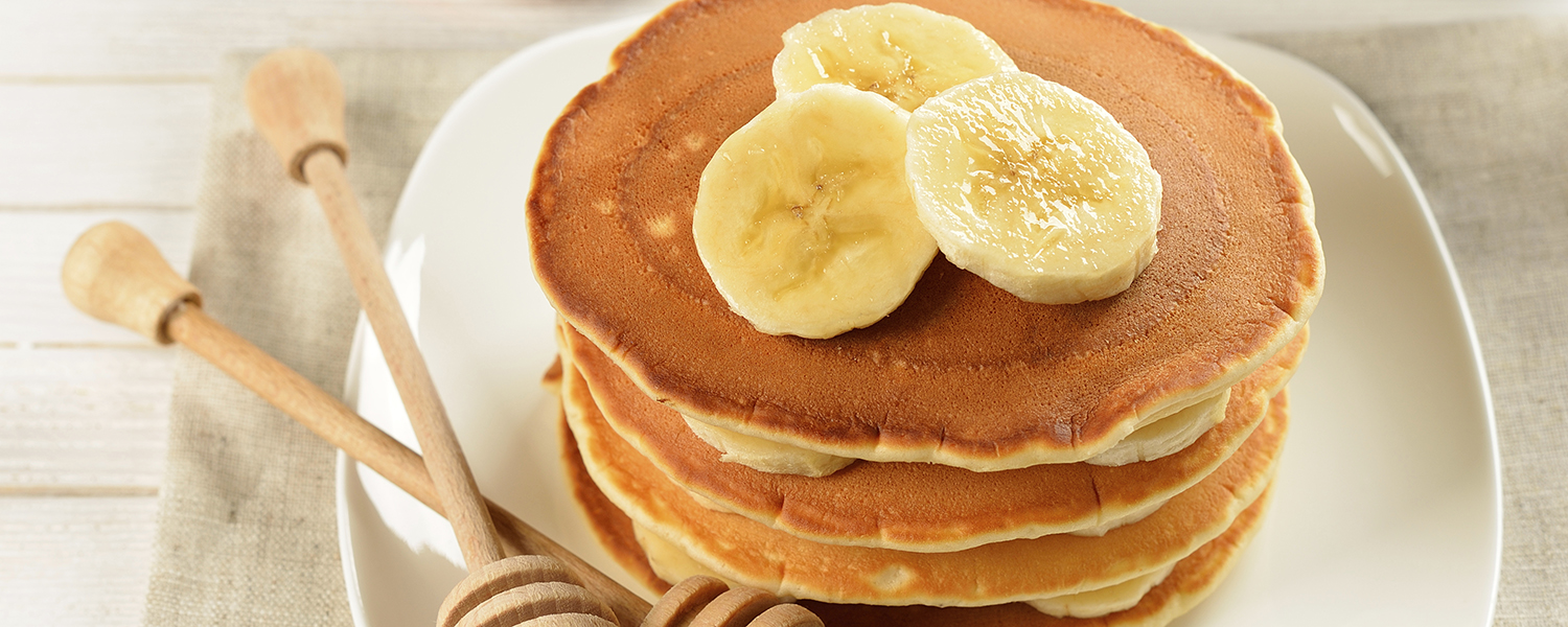 A stack of Going Bananas Pancakes on a white ceramic plate. Slices of banana top the pancakes.