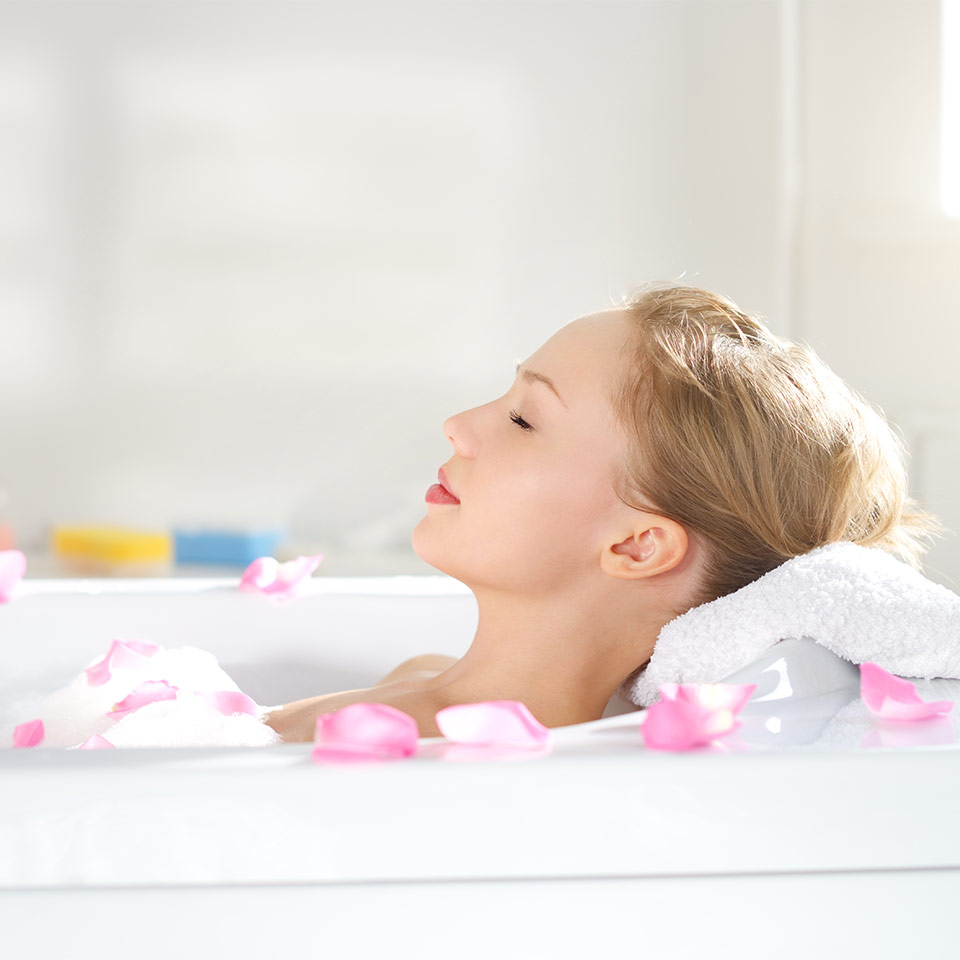 A female-presenting person with light skin and blonde hair relaxes in a bath.