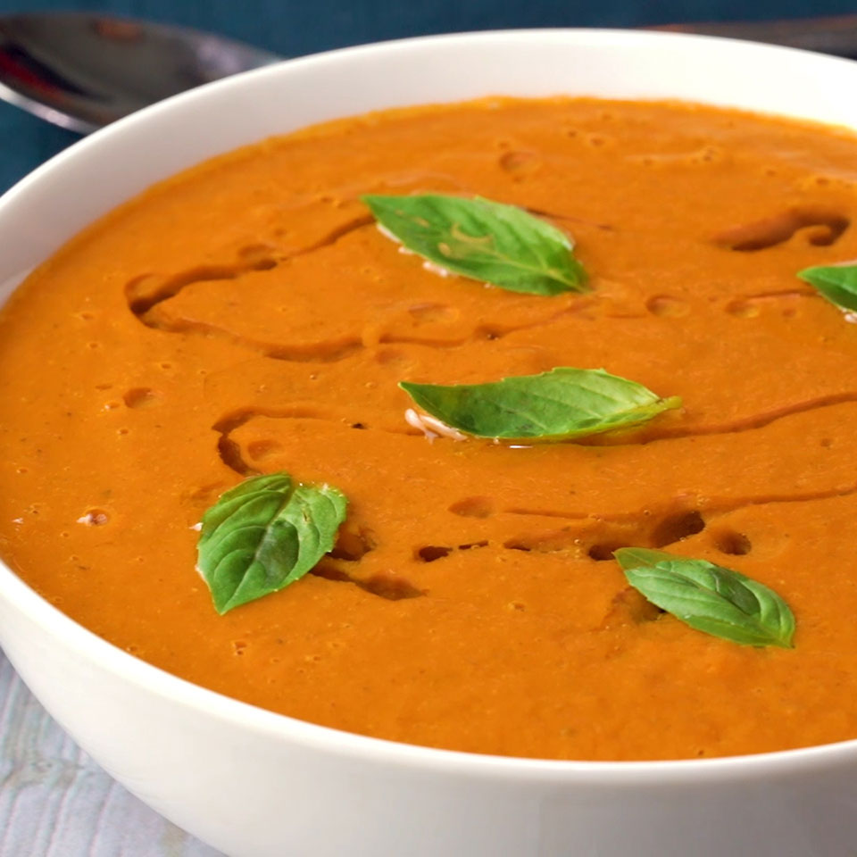 White ceramic round bowl of tomato soup dark pumpkin orange in color with a drizzle of coconut oil and five small basil leaves on top.