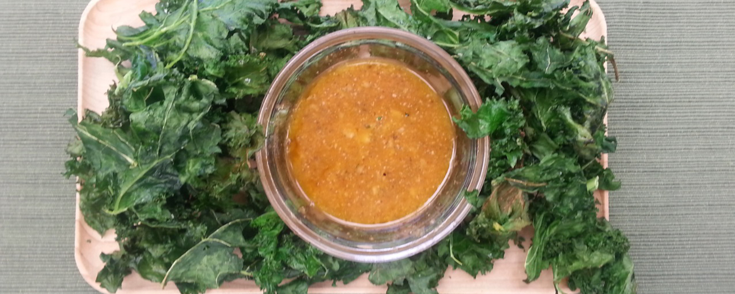 A wooden plate holding servings of kale. A dipping bowl with Siracha and Garlic Aioli at the center.