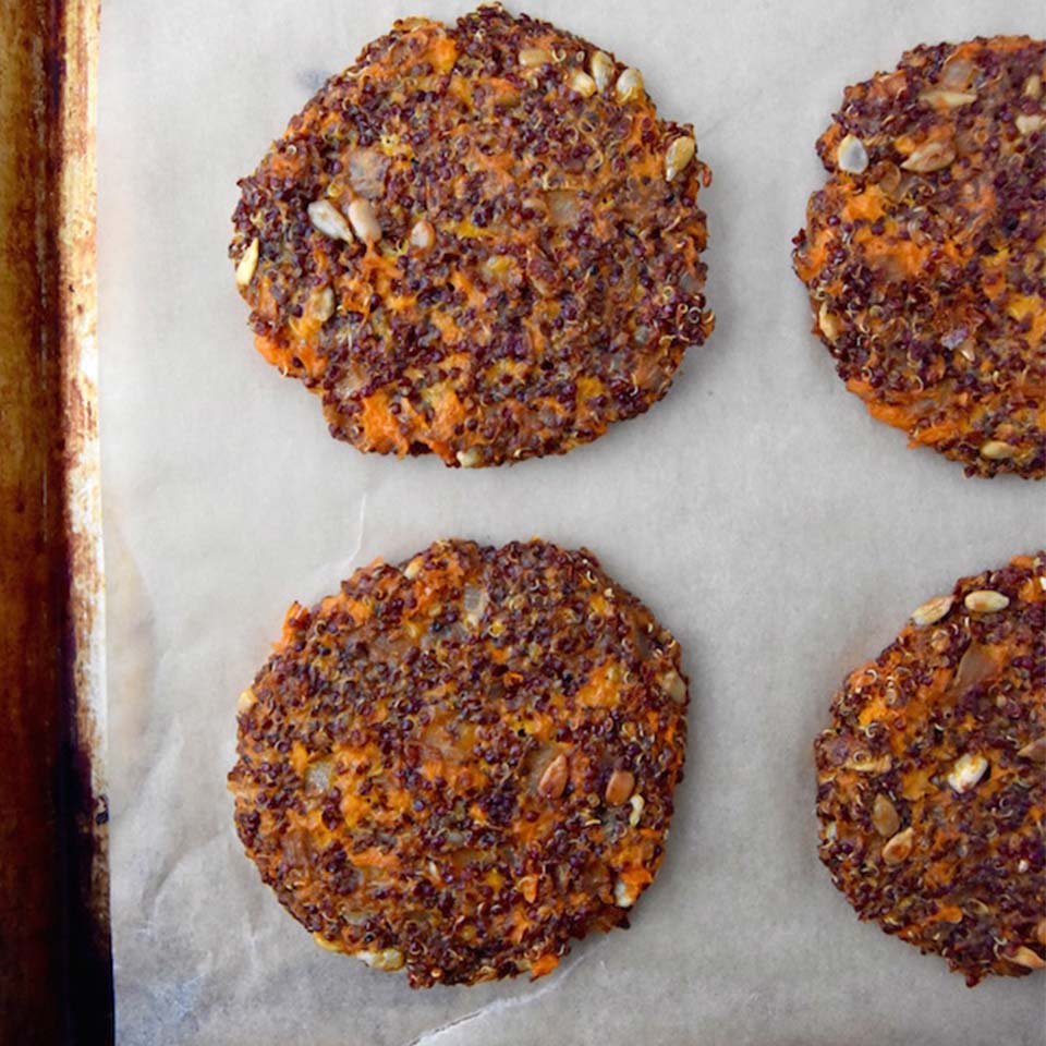 Four prepared  round flat veggie burgers on a white surface laying in a square formation with some space in between. In the mixture you can see the sweet potato color and quinoa color and some whole sunflower seeds.