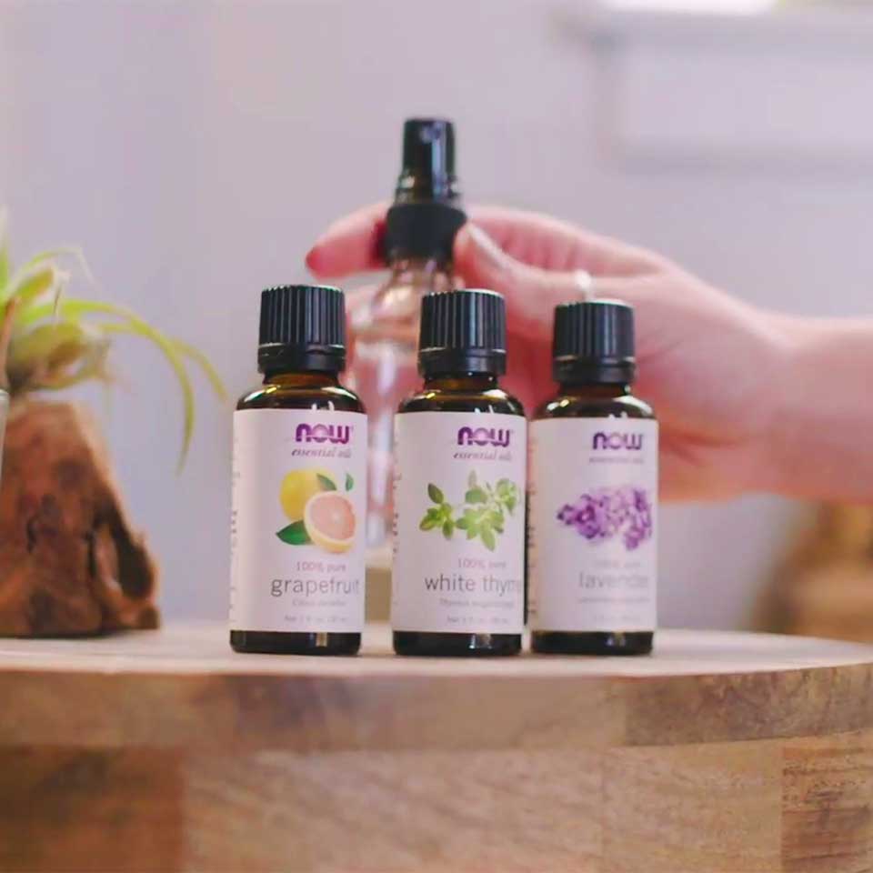 Three bottles of NOW essential oils of grapefruit, white thyme and lavender are side by side on a table with a light colored female presenting hand holding a clear glass spray bottle behind the oils. The oils are on a light wooden table and a small plant is off to the left with a white background.