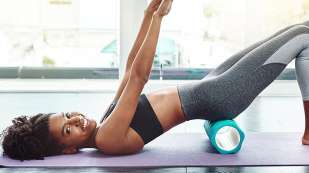 presents as woman in yoga pants and black top laying on her back with bottom raised on a foam roller and arms raised upward