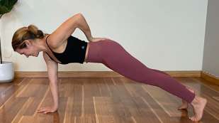 female presenting wearing burgundy yoga pants and black sports top in one arm plank position on wood floor