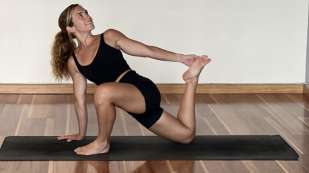 Presenting as woman in black workout shorts and top performing a yoga position on a black mat in a white room