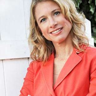 image of wendy bazilian standing in front of white fence with her arms crossed wearing a coral colored shirt
