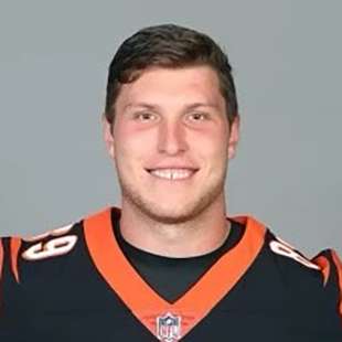 Drew Sample is smiling in this headshot photo. He has light skin and an oval shaped face with a prominent square jaw and has brown eyes and short straight dark brown hair parted to the right. He is wearing a dark brown Bengals jersey with orange trim at the v neck and the number sixty nine in white on each shoulder.