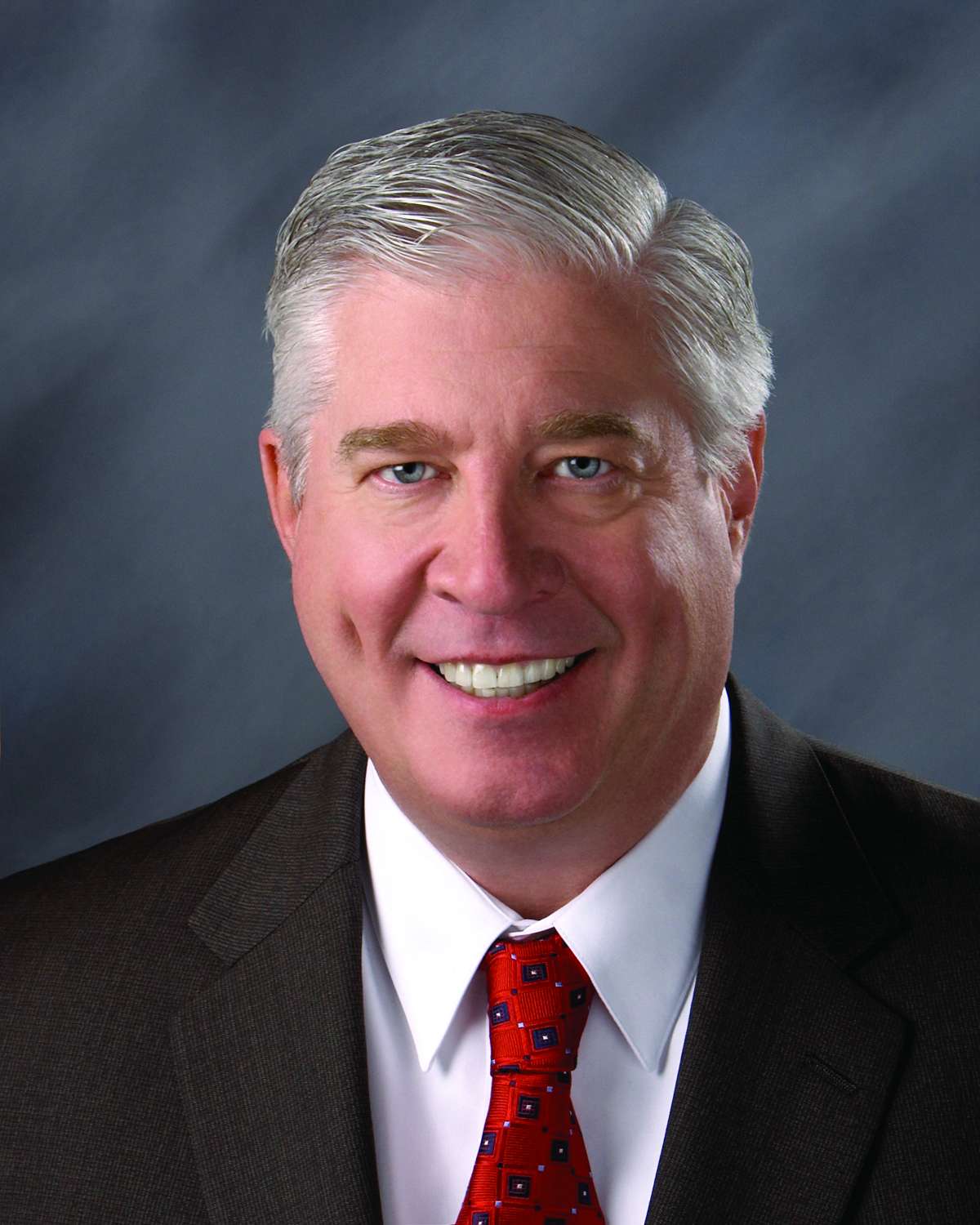 headshot image of Jim Emme, a white older male, smiling in a suit and red tie