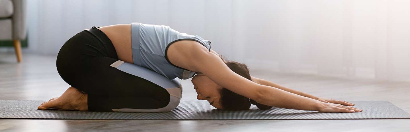 white female facing person doing a child's pose stretch with a white background wearing black pants and blue top