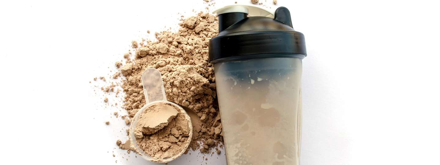 protein powder on table, in scoop with a shaker bottle next to it filled with protein powder drink