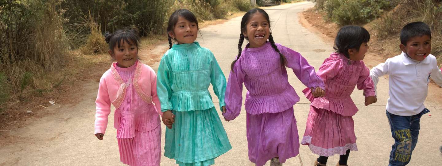 group of dark skinned children in colorful clothing holding hands walking down a dirt road