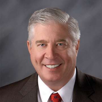 A male-presenting person with short silver hair, light skin, grey suit, and red tie smiles smiles at the camera.