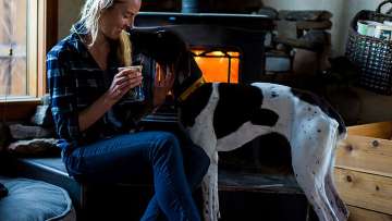 presenting as woman sitting in front of a fireplace in flannel shirt and jeans petting dog while drinking latte