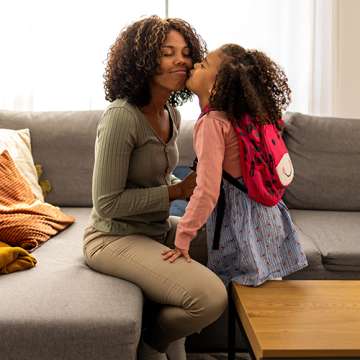 two female presenting people of dark skin tone, one presumably the mom and the other a young daughter with a backpack on hugging on a couch 